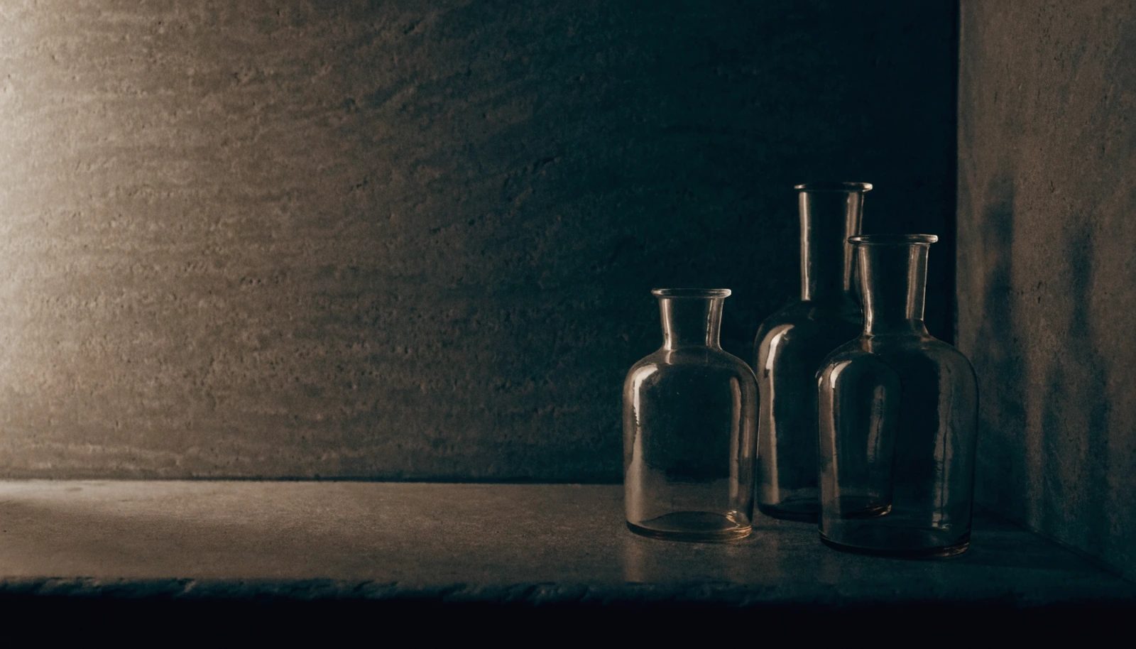 Apothecary glass vessels grouped on a dark stone bathroom shelf in first morning light.