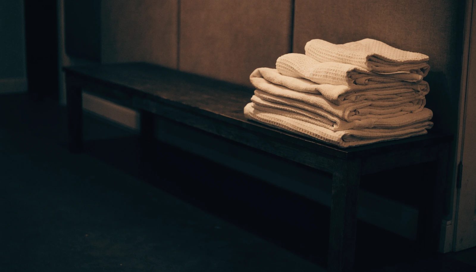 A stack of folded ivory cotton and waffle-weave towels on a dark wooden bench.